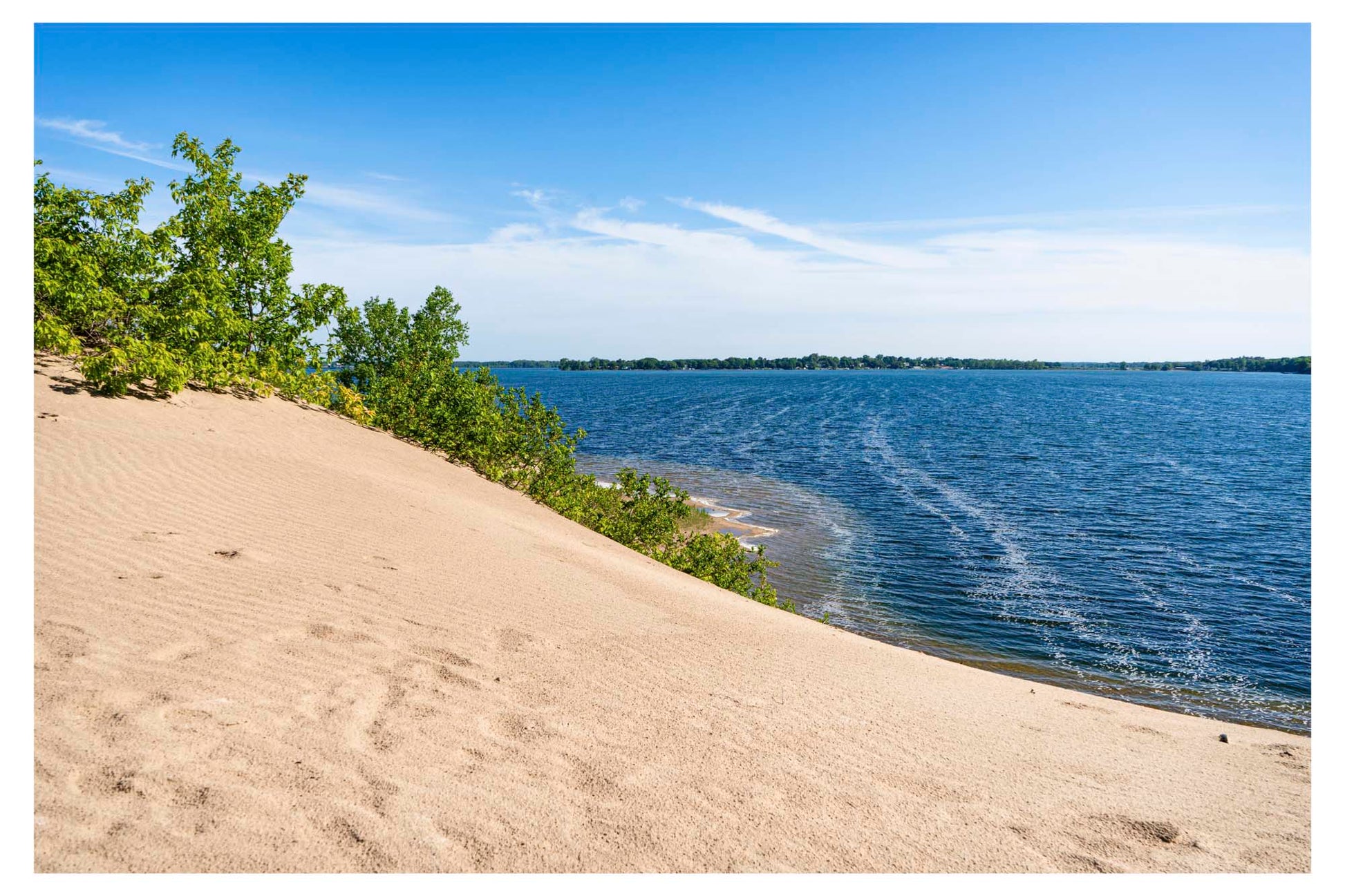 sand dune with green bush on top and water in distance.